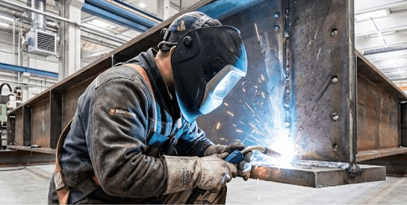Soldador industrial con equipo de protección y máscara, soldando una pesada viga de acero en una fábrica